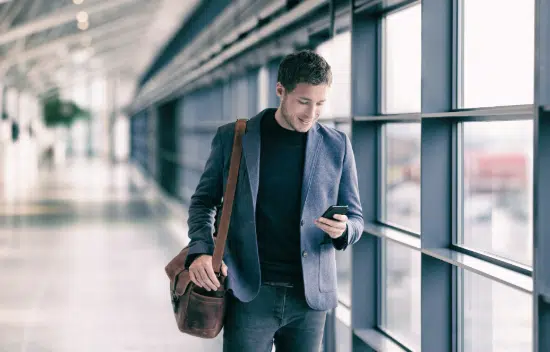 Ein Mann mit Umhängetasche nutzt sein Smartphone zur Reisekostenerfassung.