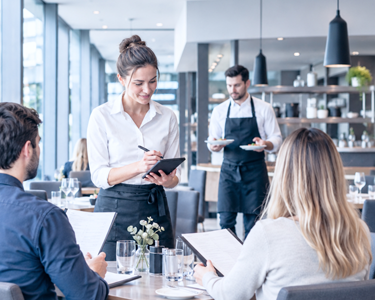 Servicekraft nimmt mit einem Tablet Bestellungen von Gästen in einem Restaurant auf, während ein Kollege Speisen serviert.